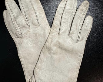May include: A pair of off-white leather gloves. The gloves are laid out on a dark surface. The gloves have a classic design with a gathered wrist detail. The leather shows signs of wear, with subtle discoloration.