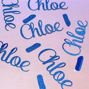 Op de afbeelding: Blauwe glitter uitsparingen van de naam "Chloe" in een cursief lettertype, verspreid over een lichtroze oppervlak. Er zijn ook verschillende blauwe glitteraccenten aanwezig. Het algehele effect is decoratief en opvallend.