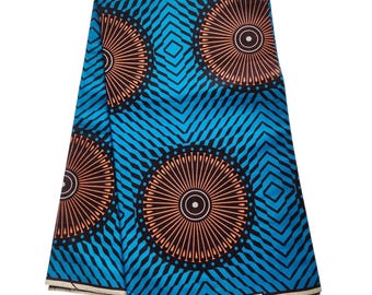 May include: A folded piece of fabric featuring a vibrant turquoise blue background with a repeating pattern of large, dark brown circular sunburst motifs and black diagonal lines. The bottom edge displays the text Veritable Premier Mumtex Wax.