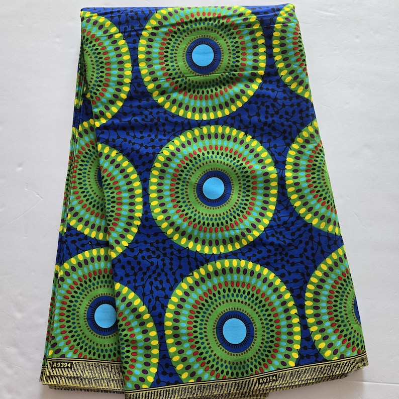 May include: A blue and green fabric with a repeating pattern of concentric circles. The circles are made up of yellow, green, red, and blue dots. The fabric has a woven texture and is folded in half.