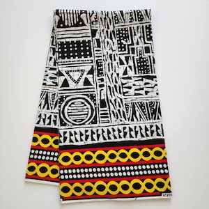 May include: Folded fabric with a bold black and white geometric pattern. The design includes triangles, circles, and rectangular shapes. The bottom edge features a border of yellow circles, red lines, and white dots. The fabric is likely cotton or a similar material.
