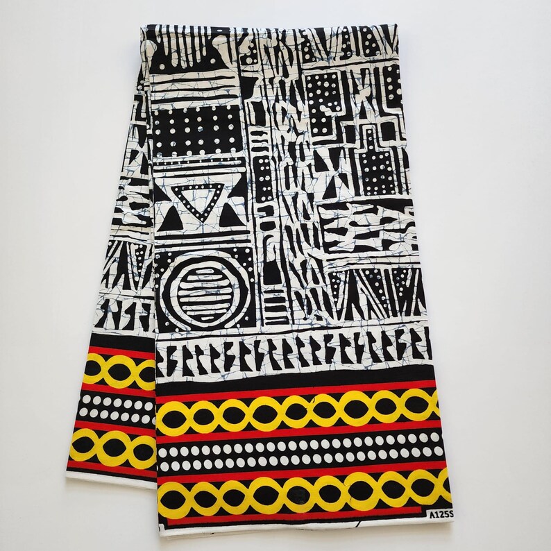 May include: Folded fabric with a bold black and white geometric pattern. The design includes triangles, circles, and rectangular shapes. The bottom edge features a border of yellow circles, red lines, and white dots. The fabric is likely cotton or a similar material.