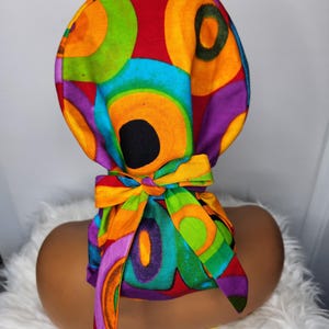 May include: A colorful surgical cap with a tie back. The cap features a pattern of overlapping circles in shades of red, orange, yellow, green, blue, purple, and black.