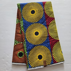 May include: A colourful fabric with a repeating pattern of yellow, blue, red and orange circles with black centres. The circles have a concentric pattern of small black dots. The fabric has the text "WAX BLOCK PRINTS HITARCET 7991" printed on the bottom edge.