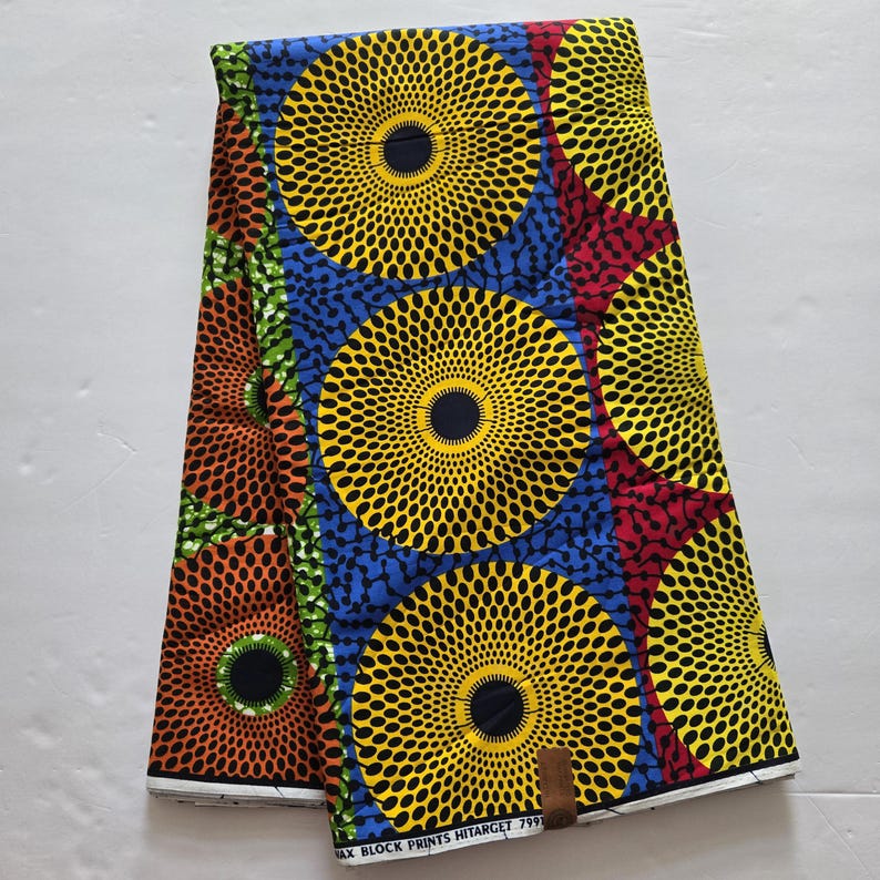 May include: A colourful fabric with a repeating pattern of yellow, blue, red and orange circles with black centres. The circles have a concentric pattern of small black dots. The fabric has the text "WAX BLOCK PRINTS HITARCET 7991" printed on the bottom edge.