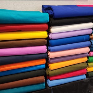 May include: Stacks of folded fabric in various colors, including teal, red, yellow, brown, and purple. Other colors include blue, black, white, pink, orange, and green. The fabric is neatly arranged, showcasing a variety of hues.