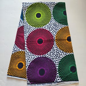 May include: White fabric with a repeating pattern of colourful circles. Each circle has a black centre and a radiating pattern of smaller circles. The colours of the circles are green, pink, purple, orange, and blue.