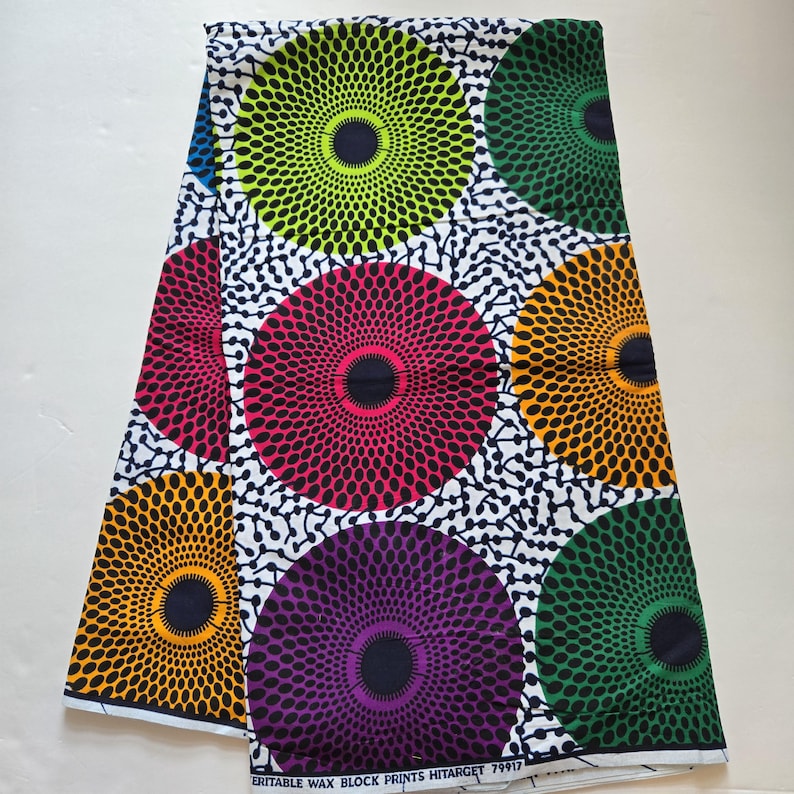 May include: White fabric with a repeating pattern of colourful circles. Each circle has a black centre and a radiating pattern of smaller circles. The colours of the circles are green, pink, purple, orange, and blue.