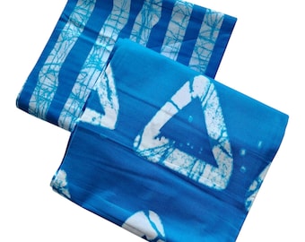 May include: Two folded blue batik fabric pieces. One piece has a striped pattern with white accents, and the other features a triangle design with white accents. The fabric has a vibrant blue color.