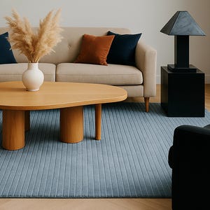 May include: A modern living room scene featuring a beige sofa with navy and rust-colored throw pillows. A wooden coffee table with a unique shape sits on a blue rug. A black lamp and side table are on the right, and a floor lamp is on the left.
