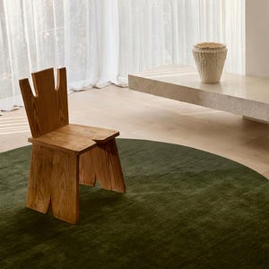 May include: A wooden chair with a unique cutout design stands on a dark green rug. The chair is made of light-colored wood. A textured, off-white vase sits on a stone shelf. Sheer white curtains are in the background.