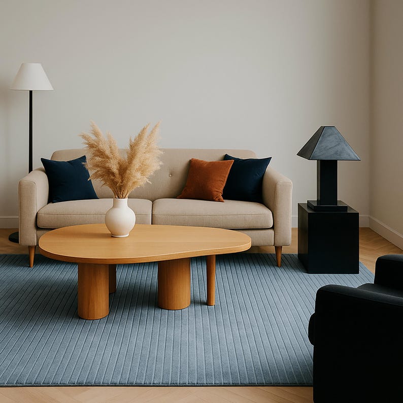 May include: A modern living room scene featuring a beige sofa with navy and rust-colored throw pillows. A wooden coffee table with a unique shape sits on a blue rug. A black lamp and side table are on the right, and a floor lamp is on the left.