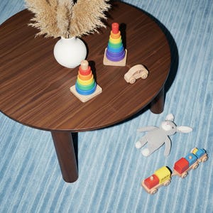 May include: A wooden table displays children's toys, including two rainbow stacking rings, a wooden car, a gray stuffed bunny, and a colorful wooden train. A white vase with dried plants sits on the table. The table is set on a blue rug.