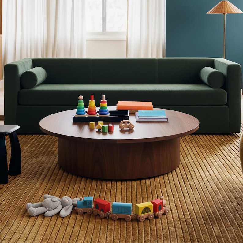 May include: A living room scene featuring a dark green sofa with bolster pillows, a round wooden coffee table, and a mustard-colored rug. The table displays wooden toys, books, and a toy car. A wooden train and a stuffed animal are on the rug.