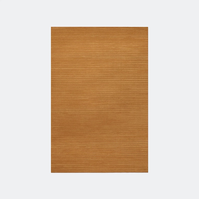 May include: A rectangular, golden-brown rug with a ribbed texture. The rug's surface is covered in horizontal lines, creating a textured appearance. The rug is a solid color and appears to be made of a soft material.