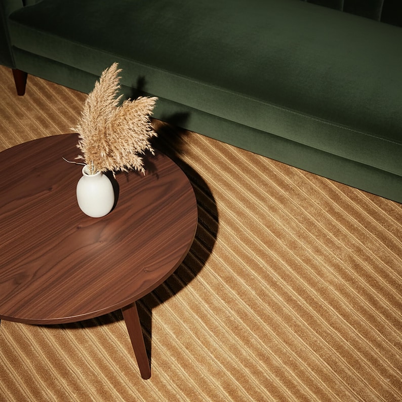 May include: A round, dark brown wooden coffee table with three angled legs. A small white vase holds dried ornamental grass. The table sits on a striped, tan rug, with a dark green velvet sofa in the background.