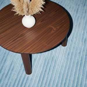 May include: A round, dark brown wooden coffee table with three legs. A white vase with dried ornamental grass sits on the table. The table is on a light blue, textured rug.