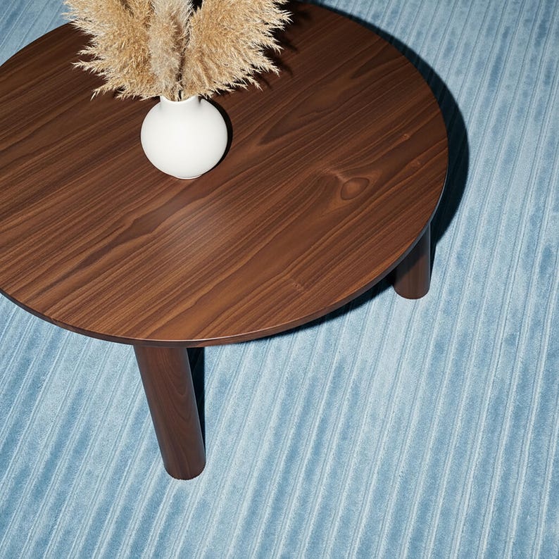 May include: A round, dark brown wooden coffee table with three legs. A white vase with dried ornamental grass sits on the table. The table is on a light blue, textured rug.