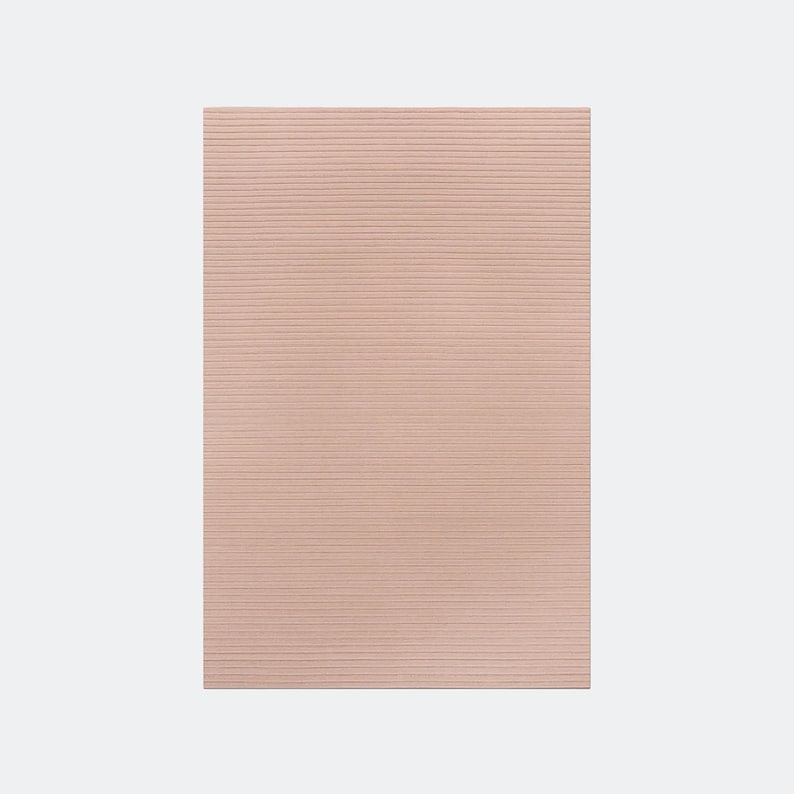May include: A rectangular, blush-colored rug with a ribbed texture. The rug's surface is covered in horizontal lines, creating a subtle pattern. The rug is a solid color and appears to be made of a soft material.