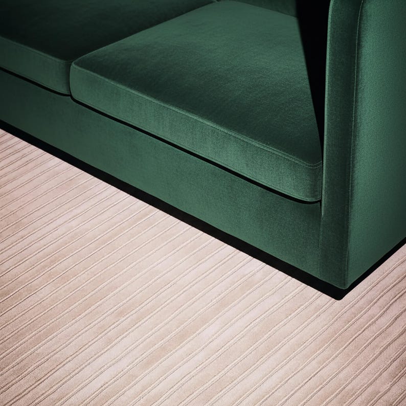 May include: A section of a modern, emerald green velvet sofa. The sofa has a low profile and a minimalist design. The rug beneath the sofa is a light beige color with a ribbed texture, adding a subtle contrast to the rich green.
