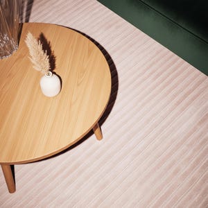 May include: A round wooden coffee table with three legs, a small white vase with dried ornamental grass, and a section of a green velvet sofa. The rug is a light pink color with a ribbed pattern.