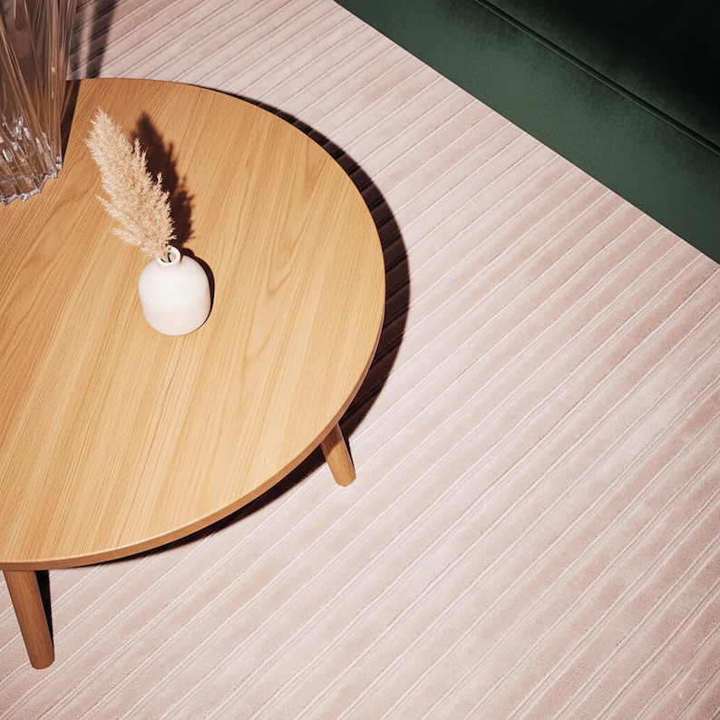 May include: A round wooden coffee table with three legs, a small white vase with dried ornamental grass, and a section of a green velvet sofa. The rug is a light pink color with a ribbed pattern.