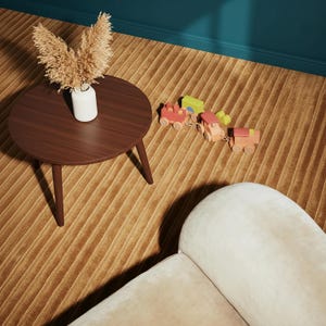 May include: A dark brown, round coffee table with three legs, a white vase with dried flowers, and a wooden toy train set on a textured, golden-brown rug. A cream-colored sofa is in the foreground, and the wall is teal.