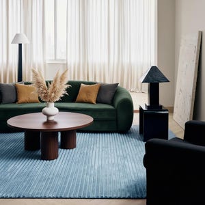 May include: A modern living room with a green velvet sofa, a round wooden coffee table, and a light blue rug. Decorative pillows in shades of gray and gold accent the sofa. A black lamp and armchair complete the stylish interior.