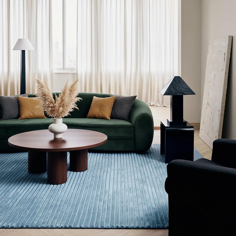 May include: A modern living room with a green velvet sofa, a round wooden coffee table, and a light blue rug. Decorative pillows in shades of gray and gold accent the sofa. A black lamp and armchair complete the stylish interior.