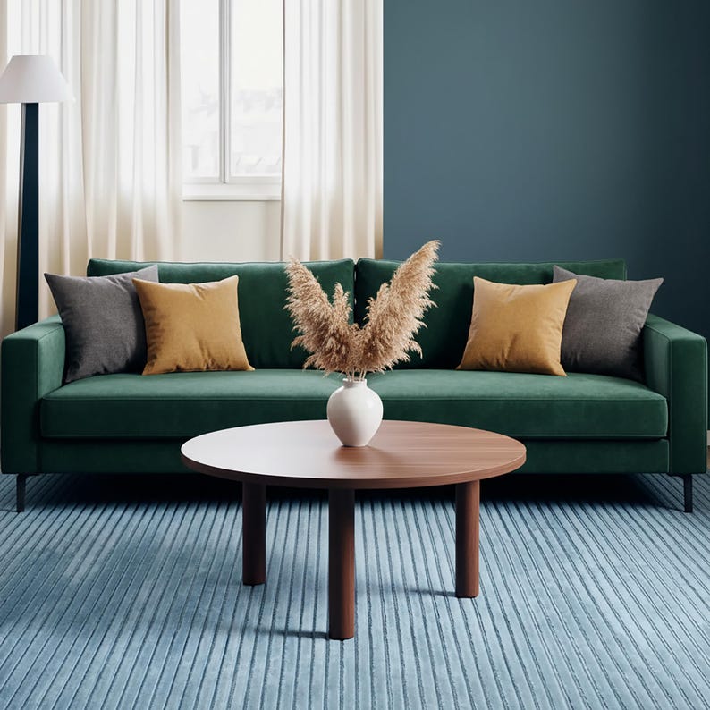 May include: A green velvet sofa with gray and gold throw pillows. A round wooden coffee table sits on a blue striped rug. A white vase holds dried plants. The room has a modern, minimalist aesthetic.
