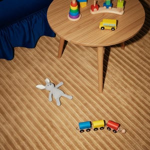 May include: A wooden round table with toys, including a yellow school bus, a train, and a colorful stacking ring toy. A gray stuffed bunny lies on the tan striped carpet. A blue skirted piece of furniture is in the background.