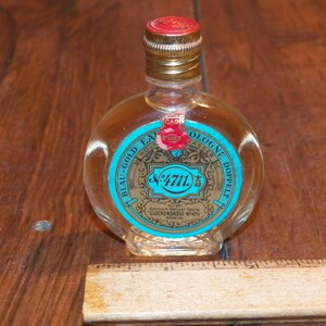 May include: A clear glass bottle of 4711 Blau-Gold Eau de Cologne with a gold-colored screw cap and a red seal on the neck. The label is turquoise and gold with black text. A wooden ruler is visible at the base, showing a width of 3 inches.