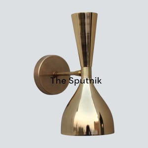 Peut inclure: Une applique murale dorée avec un abat-jour évasé en forme de cloche et un sommet fin et pointu. L'applique est montée sur une base circulaire et présente un design simple et élégant. Le texte "The Sputnik" est visible sur l'applique.