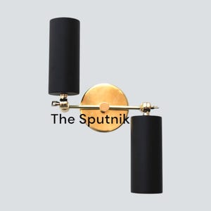 Puede incluir: Una lámpara de pared dorada con dos pantallas cilíndricas negras. La lámpara está etiquetada como "The Sputnik".