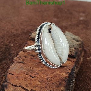Op de afbeelding: Een zilveren ring met een witte kauri schelp in een zilveren bezel. De ring heeft een getextureerde band en een gedetailleerd ontwerp.