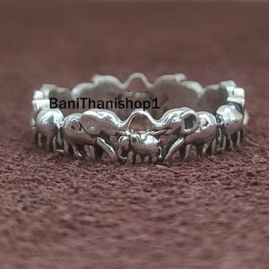 Peut inclure: Bague en argent avec un motif circulaire d'éléphants, chaque éléphant étant orienté dans la même direction, les trompes se touchant. La bague est posée sur une surface brune.