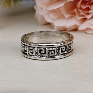 Könnte beinhalten: Ein silberfarbener Metallring mit einem durchgehenden, geprägten griechischen Mäander-Muster auf dem äußeren Band. Der Ring hat eine polierte Oberfläche mit abgedunkelten vertieften Bereichen innerhalb des geometrischen Designs für Kontrast.
