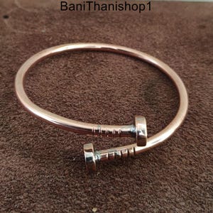 May include: A rose gold colored metal bracelet with a nail design. The nail is made of silver metal and has a textured surface.