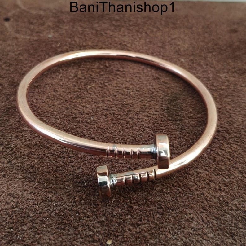 May include: A rose gold colored metal bracelet with a nail design. The nail is made of silver metal and has a textured surface.