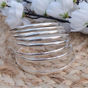 May include: A collection of silver bangles, reflecting light, sits on a textured, woven surface. White flowers with green stems are in the background. The bangles are smooth and shiny, suggesting a jewelry theme.