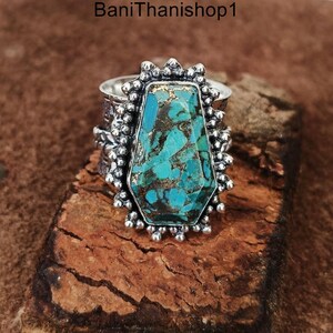 May include: A silver ring with a large turquoise gemstone set in a geometric shape. The ring has a textured band and a decorative border around the stone.