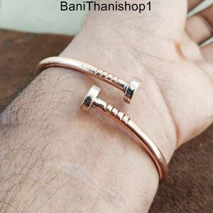 May include: A gold-toned bangle bracelet with a nail design. The bracelet is open and has a simple, minimalist style.