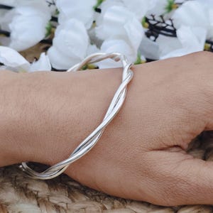 Könnte beinhalten: Ein silberfarbenes Armband aus verdrehtem Draht. Das Armband wird am Handgelenk getragen, mit einer gewebten Oberfläche im Hintergrund. Weiße Blumen sind im Hintergrund zu sehen.