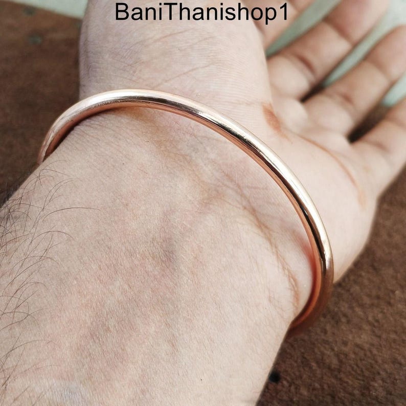 May include: A simple, polished copper bangle bracelet. The bracelet is round and smooth, with a shiny finish.