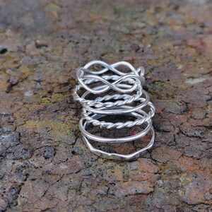 May include: A stack of silver rings with various designs, including infinity symbols and twisted bands. The rings are displayed on a textured, brown surface, showcasing their intricate details and reflective qualities. The rings are a mix of simple and ornate styles.