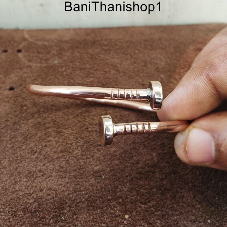 May include: A copper bracelet with two nail-shaped accents. The bracelet is open and has a hammered finish.