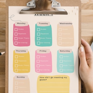 May include: A weekly music practice planner on a clipboard with a metal clip. The sheet features colourful daily sections for Monday through Sunday, each with checkboxes for Scales, Repertoire, Music Theory, and Ear training, plus a goal review section.