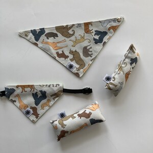 Puede incluir: Un conjunto de accesorios para mascotas, que incluye dos pañuelos triangulares, un collar y dos juguetes rectangulares. Los artículos son blancos con un patrón repetido de varios animales de safari en marrón, beige y gris. Cada artículo tiene una pequeña etiqueta con un logotipo circular.