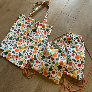 Op de afbeelding: Drie herbruikbare boodschappentassen met een witte achtergrond en een kleurrijk patroon van fruit, groenten en kruiden. De tassen zijn gemaakt van katoen en hebben oranje trekkoorden.