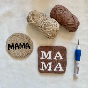 May include: Two balls of yarn, one beige and one brown, are next to two punch needle projects. One project is a round rug with the word "MAMA" in black lettering. The other project is a square rug with the word "MAMA" in white lettering. A blue punch needle tool is also included.